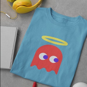 May include: A light blue t-shirt featuring a red Pac-Man ghost wearing a yellow halo. The ghost has white eyes with purple pupils.