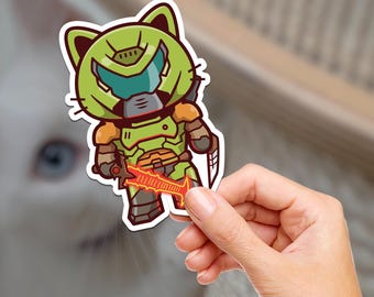 Pode incluir: Um adesivo com um gato de desenho animado em uma armadura verde e laranja, segurando uma espada vermelha brilhante. O gato tem um capacete com uma viseira azul. O adesivo é segurado por uma mão.