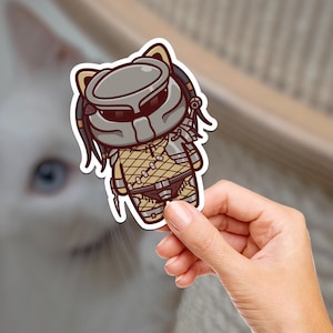 May include: A sticker of a cartoon cat dressed as a Predator. The cat is wearing a grey and black mask with red accents, a brown and tan camouflage suit, and a black belt. The sticker is on a white background.