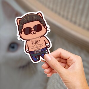 May include: A cartoon sticker of a cat wearing sunglasses and a wrestling outfit. The cat has a muscular body and a tattoo that says "Kitty Cage".