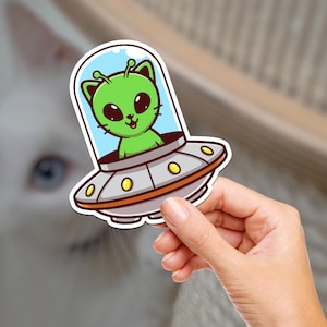 May include: A green alien cat wearing a space helmet is flying in a silver and orange spaceship. The spaceship has a round window and three yellow lights.