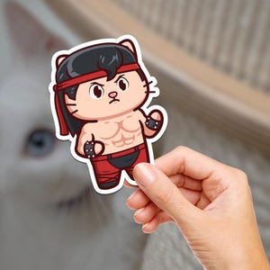 May include: A cartoon sticker of a cat dressed as a wrestler. The cat is wearing a red headband, red shorts, and black wristbands. The cat has a determined expression on its face.