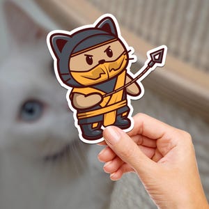 May include: A die-cut sticker featuring a cartoon cat dressed as a ninja, with a yellow mask and outfit, holding a spear. The cat is brown with black accents and a black hood. The sticker is held by a hand.