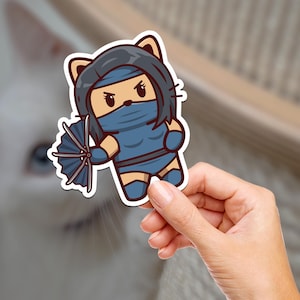 Puede incluir: Una pegatina de dibujos animados azul y blanca de un gato vestido como un guerrero ninja sosteniendo un abanico. El gato tiene el pelo negro y una máscara azul.