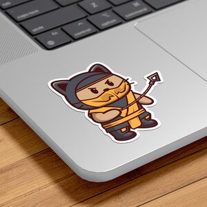 May include: A sticker featuring a cartoon cat dressed as a ninja, with a yellow mask and outfit, and a whip. The cat is brown and tan, with black boots and a black hood. The sticker is on a laptop.