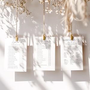 May include: Three white paper sheets with black text are suspended by gold clips and twine. The sheets have different text, including checklists and questions. The background is a white wall with dried flowers and shadows.