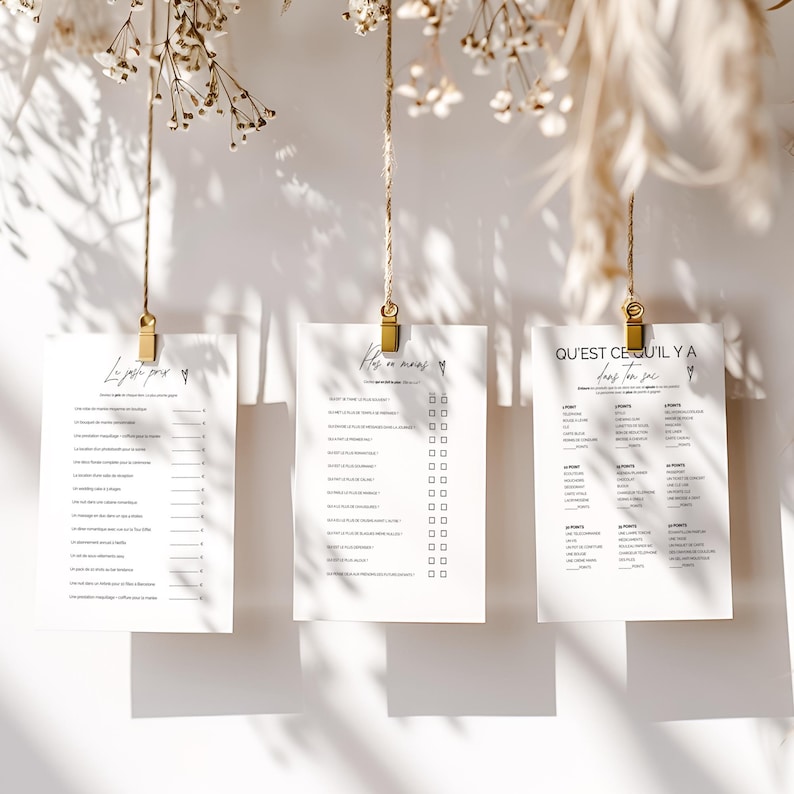 May include: Three white paper sheets with black text are suspended by gold clips and twine. The sheets have different text, including checklists and questions. The background is a white wall with dried flowers and shadows.