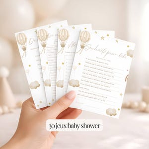 Peut inclure: Quatre cartes de jeux pour une baby shower sont tenues à la main. Chaque carte présente une illustration de montgolfière beige et dorée, avec du texte et des étoiles. Les cartes ont un fond blanc avec le texte "30 jeux baby shower" en bas.
