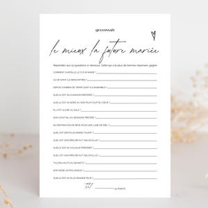 Peut inclure: Une carte de jeu blanche avec du texte noir et un cœur. Le texte manuscrit dit "Qui Connait le mieux la future mariée". La carte contient des questions à compléter sur la future mariée, avec un total de 15 questions.