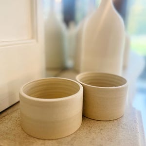 Może przedstawiać: Dwie ceramiczne miski w kolorze kremowym z lekko fakturowaną powierzchnią. Miski są okrągłe i mają prosty design.