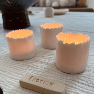 May include: Three white ceramic candle holders with a textured, uneven rim. The candles are lit and glowing. A small, rectangular, beige tile with the word "ÉMOTION" is in the foreground.