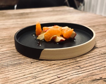 May include: A black and white ceramic plate with a small portion of orange and apple slices on top. The plate is sitting on a wooden surface.