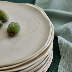 Pode incluir: Uma pilha de pratos de cerâmica esbranquiçada com bordas irregulares. Os pratos têm uma superfície sutilmente texturizada e uma borda marrom clara. Três amêndoas verdes são colocadas em cima dos pratos. O fundo inclui tecido verde.