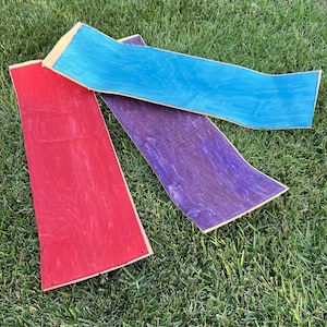 May include: Three wooden boards stained in different colors: red, purple, and blue. The boards are laid out on a green grassy surface.