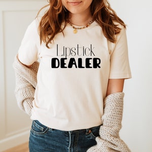 Pode incluir: Uma camiseta branca com o texto "Lipstick DEALER" impresso em preto.