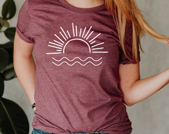 Peut inclure: Un t-shirt marron avec un motif blanc représentant un soleil couchant sur une ligne ondulée.