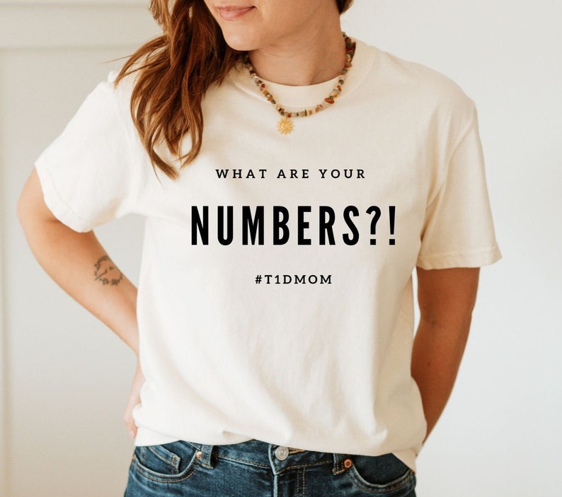 Pu&ograve; includere: Una maglietta bianca con il testo "WHAT ARE YOUR NUMBERS?!" e l'hashtag "#T1DMOM" stampato su di essa.