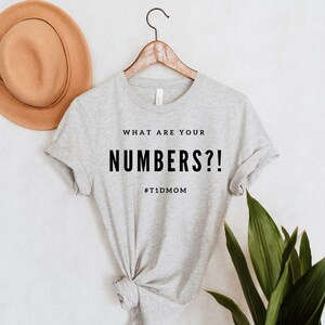 Pu&ograve; includere: Maglietta grigia con il testo "WHAT ARE YOUR NUMBERS?!" e l'hashtag "#T1DMOM" stampato su di essa.