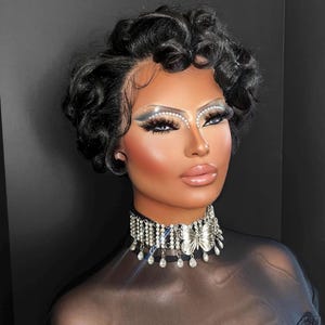 May include: A silver-toned choker necklace featuring a central butterfly motif, multiple rows of clear rhinestones, and dangling teardrop-shaped crystals. The piece is worn around the neck, showcasing a metallic finish and reflective stone accents.