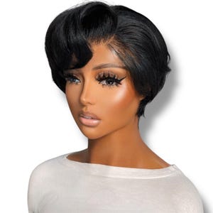 May include: A short, black, layered pixie-cut wig with a side-swept fringe and a visible lace front hairline. The hair is styled with soft volume at the crown and tapered sides, featuring a smooth, straight texture and a natural dark finish.