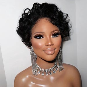 Könnte beinhalten: Eine Frau mit kurzen, lockigen schwarzen Haaren, die eine silberne Strass-Halskette und lange silberne Strass-Ohrringe trägt. Sie trägt starkes Make-up mit dramatischen Wimpern.