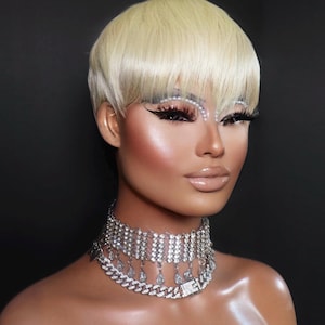 Op de afbeelding: Een meerlaagse choker-halsketting van zilverkleurig metaal met een brede band van heldere strass-steentjes, een bungelende franje van druppelvormige kristallen en een onderste schakelketting met een rechthoekige strass-sluiting.