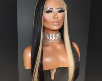 Könnte beinhalten: Eine lange, gerade Perücke mit schwarzen und blonden Strähnen. Die Perücke ist mit einem Mittelscheitel gestylt und wird auf einem Mannequin-Kopf getragen. Das Mannequin trägt eine silberne Strass-Halskette.