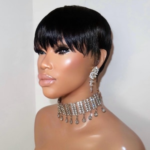 Peut inclure: Une perruque noire courte avec une frange droite et une coupe dégradée. Le mannequin porte un collier ras-du-cou argenté composé de plusieurs rangées de cristaux facettés transparents et de pendentifs en forme de goutte, avec des boucles d'oreilles assorties.