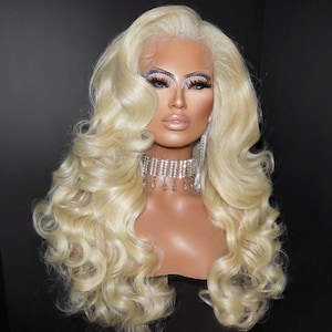 May include: A long, platinum blonde wig with voluminous, loose curls and a lace front hairline. The hair is styled with a deep side part and cascading waves that fall past the shoulders, featuring a bright, light-toned synthetic fiber.