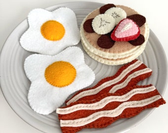 Puede incluir: Un juego de desayuno de fieltro con dos huevos blancos, un panqueque marrón con cobertura de chocolate y fresa, y tres tiras de tocino marrón en un plato blanco.