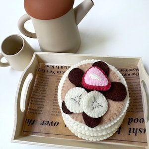 May include: A stack of felt pancakes topped with two white felt banana slices, a pink and red felt strawberry, and dark brown felt chocolate pieces, served on a wooden tray with printed paper, alongside a beige teapot and a matching cup.