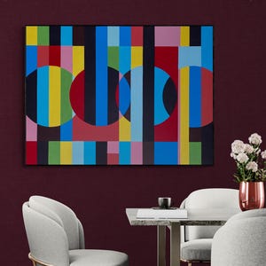 Puede incluir: Pintura geométrica abstracta con marco negro. La obra presenta formas coloridas, incluyendo círculos y rectángulos verticales en tonos de rojo, azul, amarillo y verde. La pintura cuelga sobre una mesa con sillas.