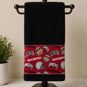 May include: A black hand towel featuring a red fabric band at the bottom with a repeating pattern of the Ohio State Buckeyes logo, the text Ohio State, and football helmets. The towel is displayed on a dark bronze metal stand.
