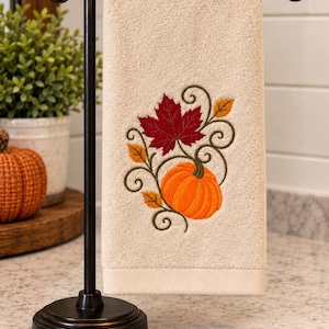 May include: A beige hand towel hanging on a dark bronze metal stand. The towel features an embroidered design of a bright orange pumpkin, a deep red maple leaf, and smaller yellow leaves, all connected by dark green swirling vine patterns.