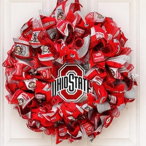 May include: A decorative wreath featuring red and white ribbons, with the words "OHIO STATE" prominently displayed in the center. The wreath is designed with a circular shape and is suspended by a red ribbon.