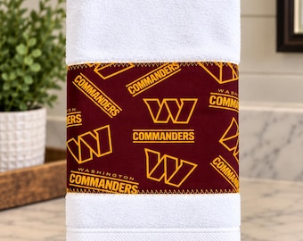May include: A white hand towel featuring a central burgundy fabric band with a repeating yellow pattern of the Washington Commanders logo and text. The towel is displayed on a black metal stand with a circular base and horizontal bar.