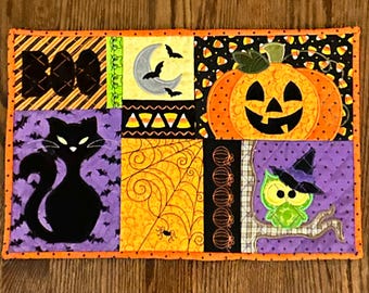 May include: A Halloween-themed quilted placemat with various appliqued designs. Features a black cat, jack-o'-lantern, owl, spider web, and the word "BOO". The color palette includes orange, black, purple, and yellow.