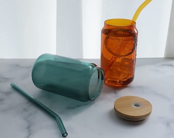 May include: Two glass can-shaped tumblers with lids and straws. One tumbler is teal green and the other is amber orange. The amber tumbler has a slice of lemon and ice cubes inside. The lids are made of wood with a silicone seal.