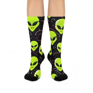 May include: Black socks with a repeating pattern of neon green alien faces. The alien heads have large black eyes and are outlined in black. Pink and white swirly lines are scattered across the black background.