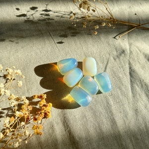 May include: A group of six iridescent, white, and blue opal tumbled stones on a light brown fabric background.