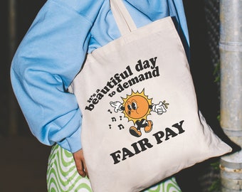 May include: A white canvas tote bag with a black and yellow graphic of a sun with the text "It's a beautiful day to demand fair pay".