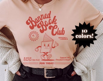 May include: A peach-colored t-shirt with a red graphic of a book character with the text "Banned Book Club Since 2023" and the text "B.Y.O.B Bring Your Own Book Join Today! Support the Freedom to Read!"  The graphic is surrounded by a sun and a starburst with the text "10 colors!"