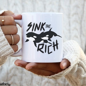 May include: White ceramic mug with a black graphic of two orcas and the text "Sink the Rich" in a bold, black font.