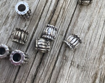 May include: A collection of small, silver beads with a ribbed design and a central hole are arranged on a weathered wooden surface. The beads have a cylindrical shape with a decorative pattern, suitable for jewellery making or crafts.