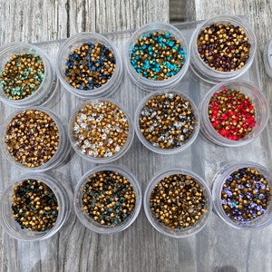 May include: Twelve small clear containers filled with various colours of rhinestones. The containers are arranged in four rows of three. The rhinestones are in shades of gold, silver, green, blue, red, and purple.