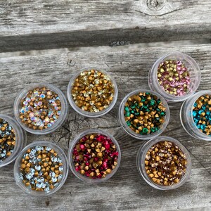 May include: Assortment of small containers filled with colourful rhinestones. The containers are clear plastic and the rhinestones are various colours, including gold, silver, red, green, and blue.