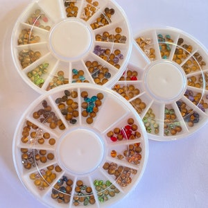 May include: Three white circular containers with compartments filled with various coloured rhinestones. Each container has 12 compartments and is designed for nail art.