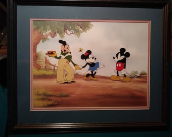 Può includere: Cel animata vintage incorniciata con Topolino, Minnie e Pippo in un ambiente pastorale. Pippo tiene un vassoio, mentre Minnie e Topolino sembrano ballare. L'opera d'arte è incorniciata con un passe-partout blu e una cornice in legno scuro.