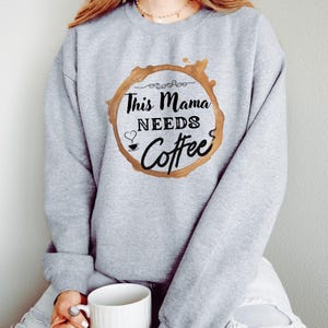 May include: A light gray sweatshirt with the text "This Mama Needs Coffee" in a brown coffee stain design. The text is in a black, handwritten style font. A white coffee mug is held in the person's hand.