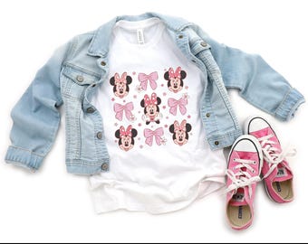 May include: A white t-shirt featuring a repeating pattern of Minnie Mouse faces and pink bows, layered under a light blue denim jacket, accompanied by a pair of pink canvas sneakers with white laces and white rubber toe caps.